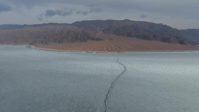 Drone footage of the frozen Orto-Tokoy Reservoir with a long ice crack across the surface, surrounded by barren mountains and shoreline in Kyrgyzstan, Central Asia