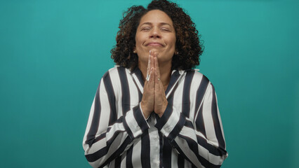 Hispanic woman with hands pressed together and eyes closed stands against teal backdrop in studio  serenity calm peace tranquility. © Krakenimages.com