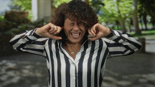 Middle aged hispanic woman in striped shirt covers ears with fingers and furrows brow beside building; irritation.