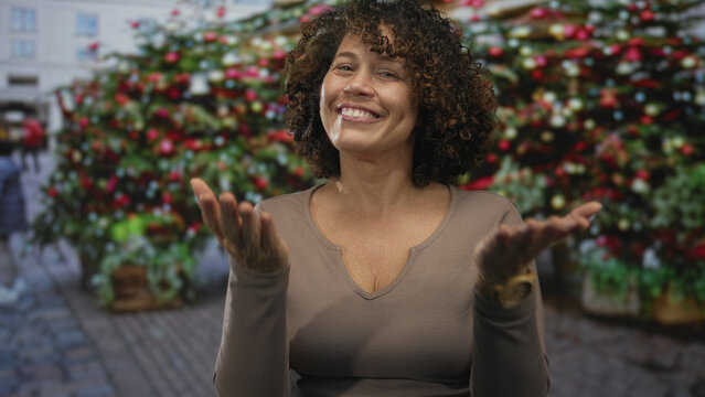 Woman beckons hand to camera on cobblestone street under decorated tree; welcome warmth anticipation joy.