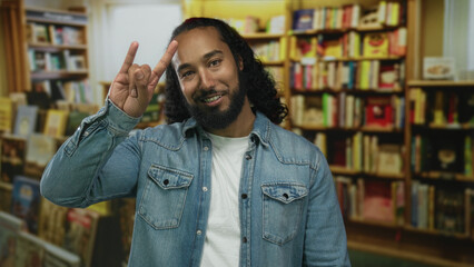 Man showing rock horns with hand in a library building, smiling amid bookshelves and wearing denim jacket and white tee  confidence. © Krakenimages.com
