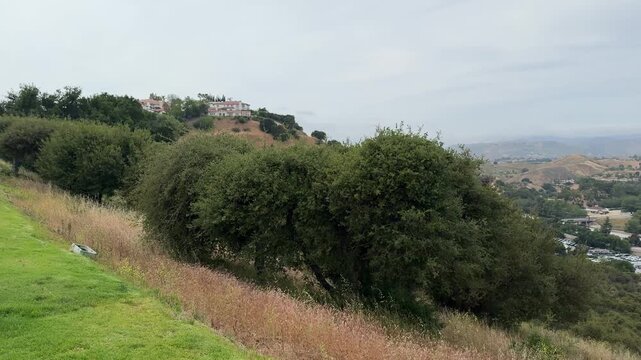 Expansive outdoor shot capturing vast valley, revealing rolling hills, dry grass, green trees, urban infrastructure, distant mountains, cloudy sky, conveying tranquil yet active environment