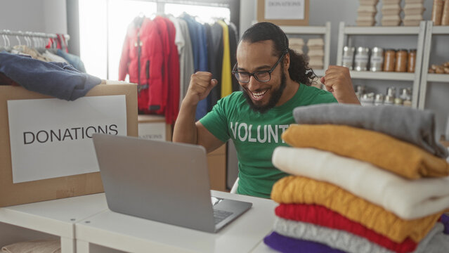 Man wearing volunteer shirt types on laptop beside donations box and stacked clothes in a building; community support joy.