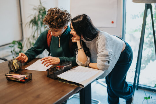 Two students lean over papers as they study and collaborate on school assignments. A casual tutoring moment shows teamwork and concentration in a bright classroom setting.