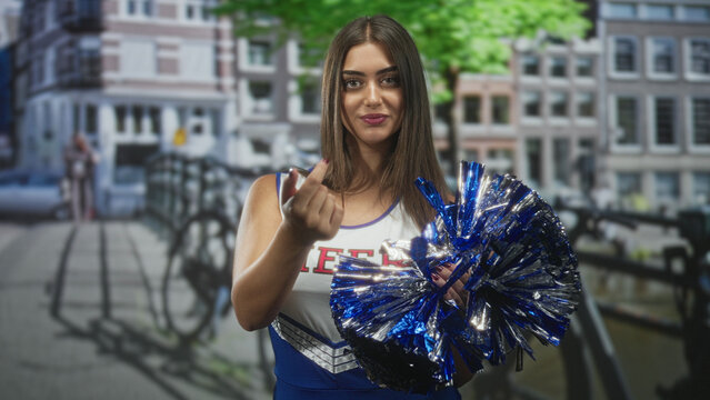 Woman cheerleader in blue uniform holding shiny metallic pompom, smiles and beckons with finger on street by canal bridge; playful confidence.