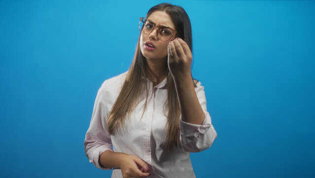 Young hispanic woman wearing glasses making a beckoning hand gesture while looking skeptical in a studio; frustration inquiry.