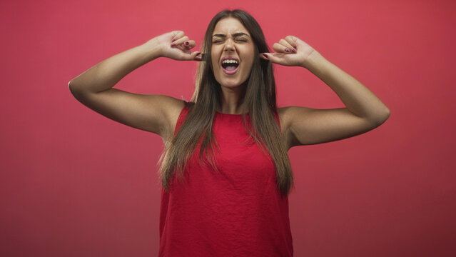 Woman in red top covering ears with fingers, arms raised and shouting in studio; frustration release.