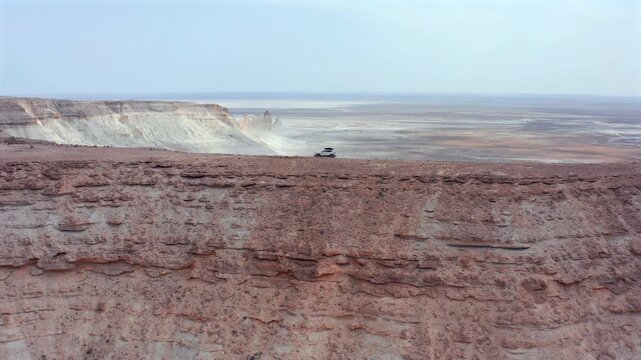 Aerial drone view of tiny SUV driving along plateau edge in Bozzhyra desert, Mangistau Kazakhstan, with vast open landscape, emphasizing scale, distance and isolation in remote arid terrain