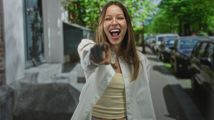 Fototapeta premium Young woman finger pointing to camera on a city street beside parked cars in daylight, wearing white shirt; confidence challenge empowerment.