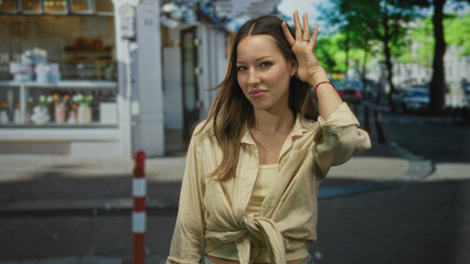 Obraz premium Woman cups hand to ear and leans forward on a busy city street near storefronts, listening to a distant sound; curiosity attention.