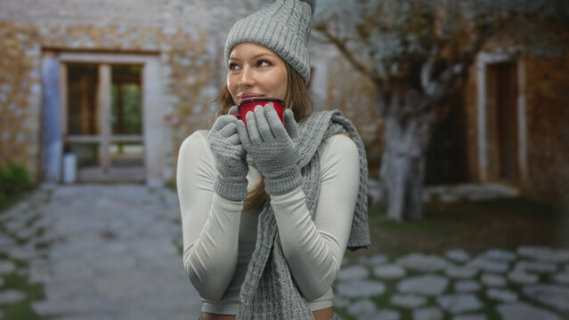 Woman holding red mug to lips with gloved hands at building entrance, wearing gray knit hat and scarf, eyes closed in winter; cozy comfort.