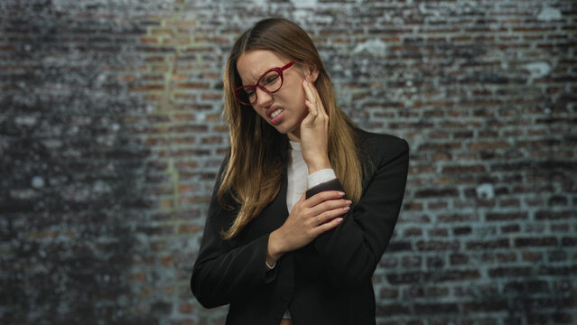 Woman holding ear with hand and clutching elbow while wincing in front of a brick building wall, wearing red glasses and black jacket; discomfort.