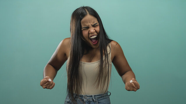 Young hispanic woman with hands raised and fists clenched screaming in a studio teal backdrop wearing tanktop and jeans; frustration release.
