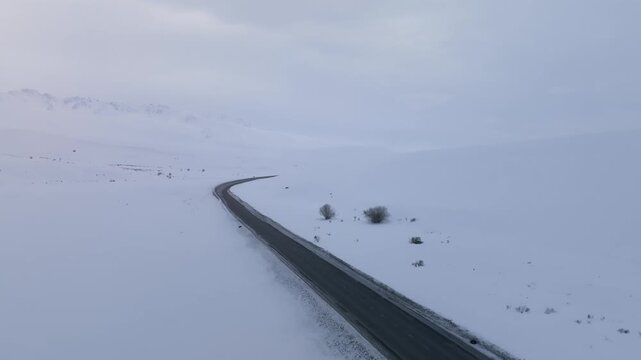 Drone footage of a winding asphalt highway cutting through deep snow at Ala-Bel Pass, Naryn Region, Kyrgyzstan, under overcast winter sky with no traffic