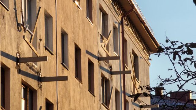 Close-up view of a worn multi-story apartment building with visible metal brackets where balconies once stood, indicating extensive renovation work at sunset.