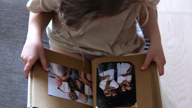 High angle view of a child sitting on the floor, holding and slowly opening a nostalgic photo album, remembering happy family moments and exploring her personal family history and roots