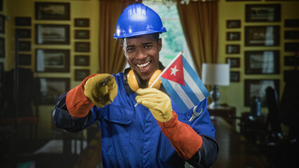 Naklejka premium Man in hardhat holding cuban flag, wearing yellow gloves and earmuffs, smiling while pointing finger toward camera in building; national pride.
