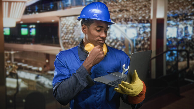 Man in hardhat wearing blue coverall and noise cancel headphones holding laptop with wrench and hand to chin in building; focused problem solving.