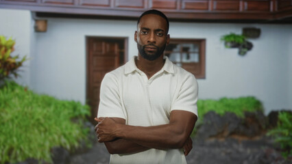 Fototapeta premium Young man with arms crossed at house entrance building, wearing white polo and visible forearms and beard; confidence.