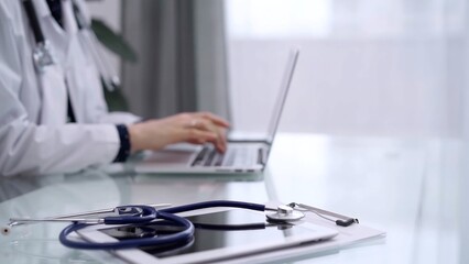 Fototapeta premium Tablet computer and blue stethoscope are on the glass table while doctor woman is using laptop computer on the glass table. Medicine concept