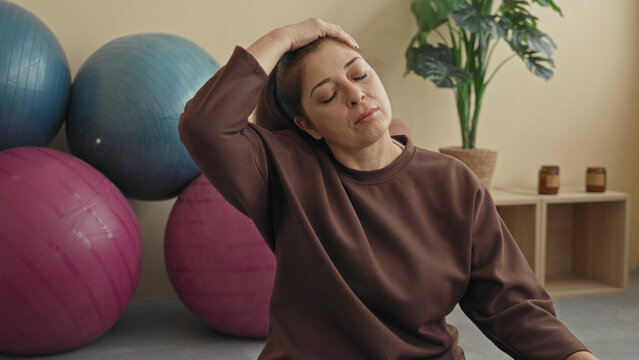 Woman doing seated neck stretch with hand on head and eyes closed, wearing brown sweatshirt, exercise balls, plant and candles in yoga studio; relief.