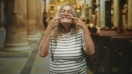 Fototapeta premium Senior woman pointing fingers to her cheeks while smiling in a church nave among pews and stone columns; joy.