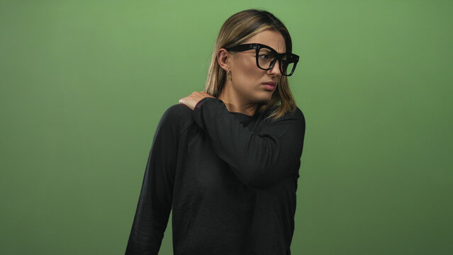 Young blonde woman holding shoulder with hand wincing from shoulder pain in studio green set wearing black top and oversized glasses; discomfort.