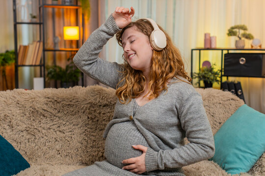 Pregnant young woman listens to music on headphones at home, gently touching her belly, dancing with careful steps and positive mood. Girl enjoys the rhythm, feeling love, connection, and happiness.