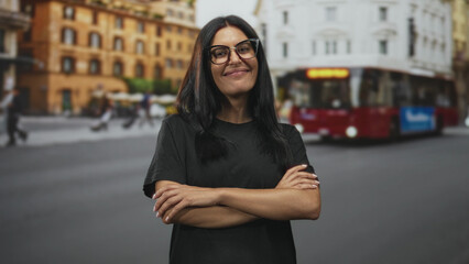 Fototapeta premium Woman with crossed arms smiling, wearing glasses, on a busy city street beside a red bus and classic buildings; confidence independence.