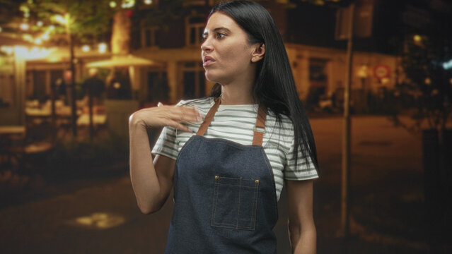 Woman in denim apron raises her hand in a dismissive gesture on street outside cafe at night near tables and lights; annoyance resilience.