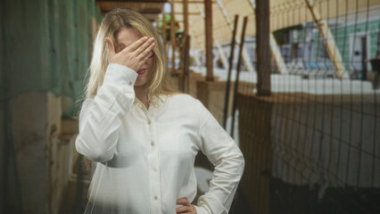 Fototapeta premium Blonde woman covers face with hand on a street platform beside a metal fence and columns, wearing white shirt, one hand on hip; shyness solitude.