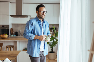 Fototapeta premium Smiling Man Enjoying Morning Coffee By Window In Modern Kitchen, Casual Home Lifestyle Moment