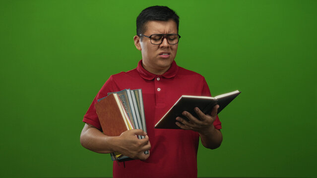 Young hispanic man holding textbooks and squinting while reading a notebook in a studio with green backdrop; study frustration.