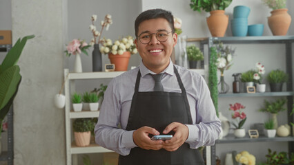 Fototapeta premium Man florist holding smartphone and smiling in studio with shelves of pots, vases and potted plants visible; contentment.