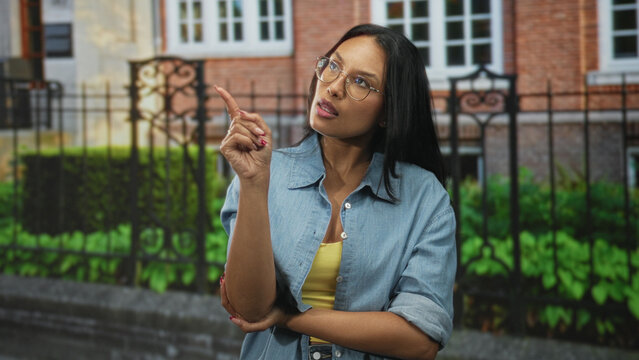 Woman points finger with raised hand and folded arm in front of building, wearing glasses and denim shirt; confusion.