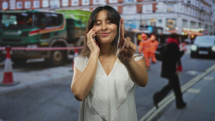 Fototapeta premium Latina woman points finger while talking on phone on a busy street construction site with cones and workers nearby; confidence resilience.