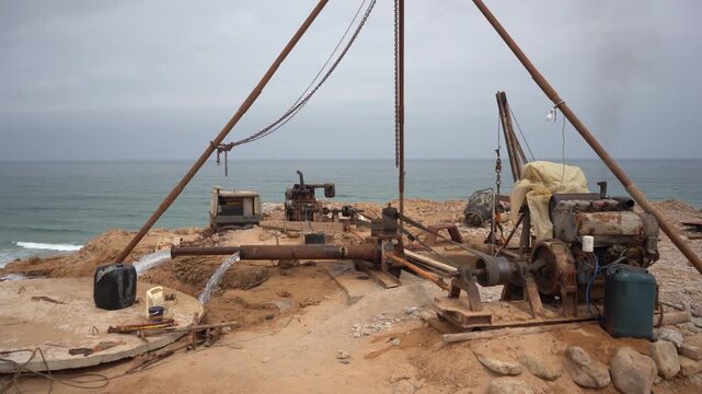 Wide shot of a rusty industrial drilling rig actively pumping water on a sandy ocean beach under an overcast sky. Features raw, authentic mechanical engine sound. Ideal for coastal engineering B-roll.