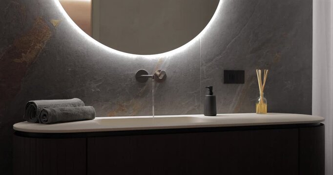 Backlit round mirror over running faucet and ribbed wood vanity, static video frames sink zone as water falls into pale basin while glowing halo washes dark stone and reveals a premium bathroom