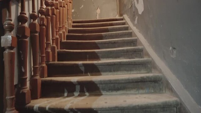 Interior view of old wooden staircase in derelict building. Steps show significant wear, surfaces covered in dust and debris. Flaking paint and plaster on walls emphasize abandonment and need of repai