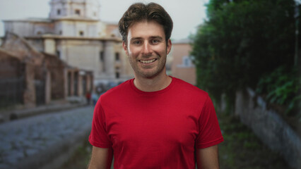 Obraz premium Man smiling with visible face and short stubble wearing a red t shirt on an old town cobblestone street near historic building; friendly happiness.