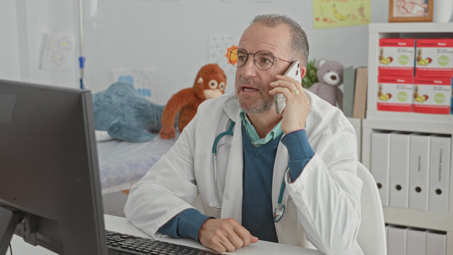 Man doctor holding smartphone to ear speaking on phone with stethoscope and computer visible in building; concerned telemedicine support.