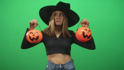 Naklejka premium Woman witch wearing wide brim hat holds two jack o lantern pumpkin buckets by raised hands showing bare midriff against green screen studio backdrop; skeptical halloween fun.