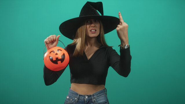 Woman in witch hat holds pumpkin bucket and shows middle finger, crop top midriff visible in studio; playful defiance.