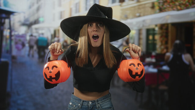 Woman wearing witch hat holds two pumpkin buckets, bare belly visible, mouth open in surprise on street; playful celebration.