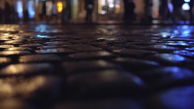 Low angle perspective captures texture of wet cobblestones on city street at night. Reflections of vibrant urban lights, a mix of warm and cool tones, illuminate pathway. Blurred silhouettes of pedest
