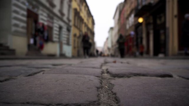 Ground level view cobblestone street. Blurry people walk urban path. Historical buildings line street. Depth of field emphasizes texture, blurring distant movement. Old European city.