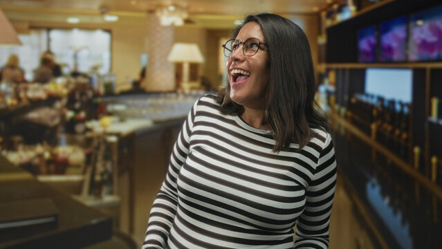 Woman wearing glasses smiling and looking up at a bar counter inside a building, striped shirt visible and relaxed posture; joy relaxed contentment.