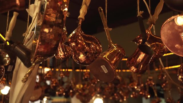 Decorative copper kitchenware, including hammered pots, pans, and cezves, suspended by rope. Warm light illuminates polished surfaces, casting reflections against dark, bokeh-filled background, evokin