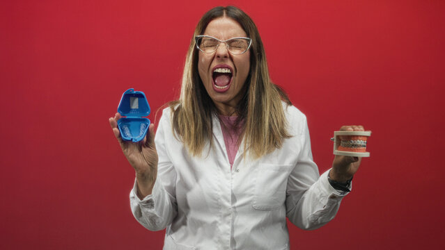 Dentist woman middle age holding clear aligner case and orthodontic dental model, mouth open wide showing teeth, wearing white coat in studio; frustration warning.