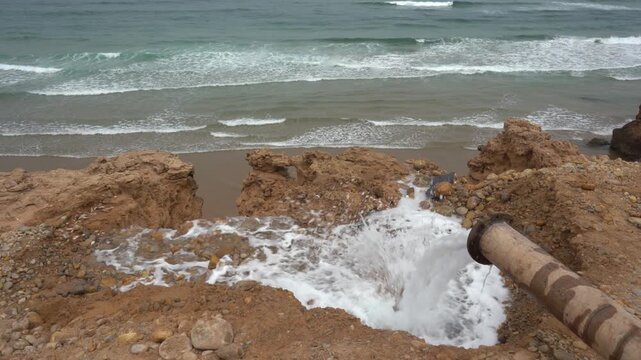High-angle shot of a rusted industrial pipe dumping foamy wastewater onto an eroded beach cliff, flowing directly into the ocean. Perfect B-roll for ocean pollution and environmental documentaries.
